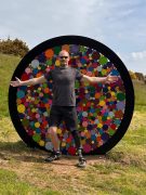 Visitor with a prosthetic leg standing in front of the multicoloured circles sculpture at St Nectan’s Glen, smiling with arms outstretched in Cornwall’s Temperate Rainforest — inclusive, accessible outdoor art installation.